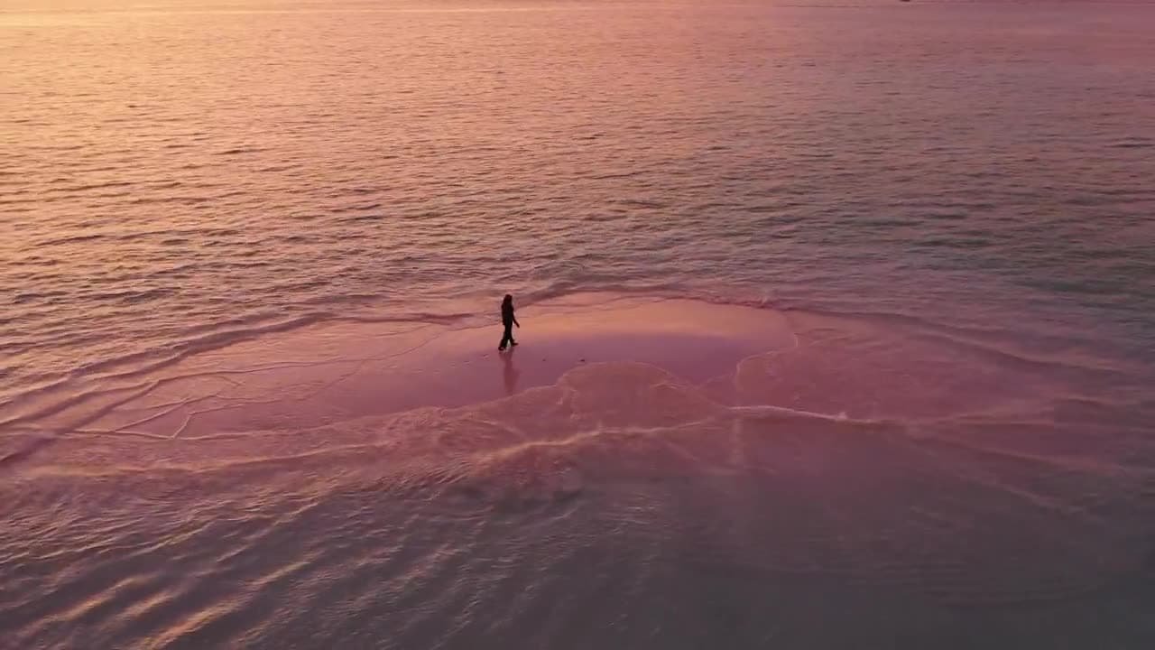 Pessoa caminhando sozinha sobre um banco de areia ao pôr do sol, com a maré indo e voltando ao redor